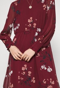 Femme portant une robe bordeaux à manches longues avec des motifs floraux roses et blancs, ainsi qu’un collier pendentif rond en or, les mains doucement jointes.