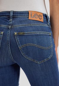 Vista trasera de una persona que lleva unos vaqueros Lee de mezclilla azul oscuro con un parche de cuero marrón visible de la marca y costuras amarillas en el bolsillo.