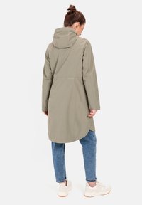 Light olive green waterproof jacket with a hood, a cinched waist, long sleeves, and a curved hem, worn over blue jeans and white sneakers.
