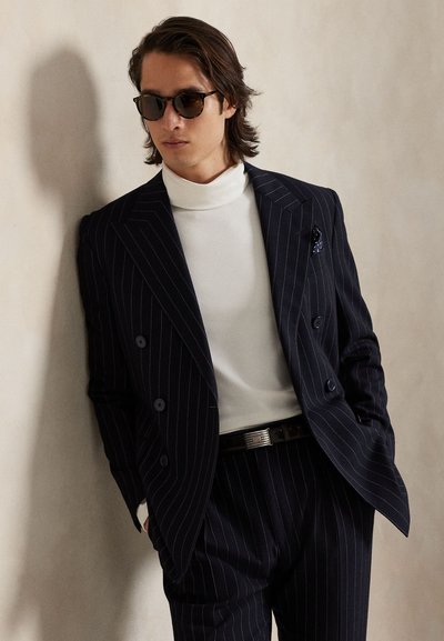 Vīrietis tumšā raibu strīpu uzvalkā, baltā turtleneck kreklā, saulesbrillēs un melnā jostā, balstās pret bēšām sienām ar rokām kabatās.
