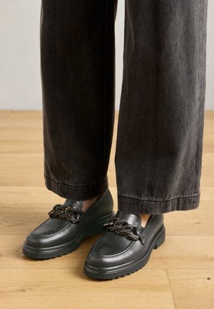 Mocassins en cuir noir avec une semelle épaisse et un accent chaîne. Assortis à un pantalon large gris foncé sur un sol en bois.