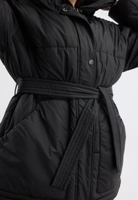 Doudoune noire avec taille ceinturée, boutons-pression, texture matelassée et poches latérales pour plus de fonctionnalité. Design moderne et minimaliste.