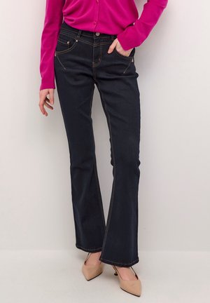 Kobieta w ciemnoniebieskich dżinsach bootcut, jasnoróżowej bluzce z długim rękawem i beżowych szpiczastych szpilkach stojąca na jednolitym białym tle.