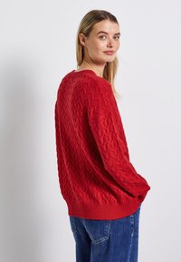 Roter gestrickter Pullover mit einem strukturierten Zickzackmuster und geripptem Saum. Lockere Passform mit überschnittenen Schultern, kombiniert mit blauen Jeans.