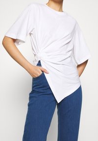 Mujer con una camiseta blanca de gran tamaño anudada en el costado, combinada con pantalones vaqueros azul de cintura alta, con la mano en el bolsillo, sobre un fondo liso.