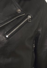 Černá kožená motocyklová bunda s stříbrnými zipy, texturovaný povrch, diagonální přední kapsa na zip a knoflíková spona na límci.