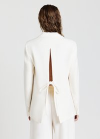 Blazer blanc avec une fente arrière profonde et une ceinture. Fabriqué en tissu lisse avec un design structuré et des manches longues.