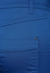 Blue fabric pants featuring a smooth texture, visible stitching, a back pocket with a metal snap, and a fitted design.