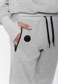 Szare dresowe spodnie wykonane z miękkiego materiału, z zamykaną na suwak kieszenią i czarną okrągłą naszywką z logo z przodu. Widoczne są czarne sznurki do regulacji.