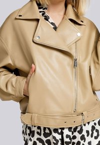 Giacca da motociclista in pelle beige con ampio colletto, zip argentate e vita con cintura. Il materiale è liscio e con una finitura opaca.