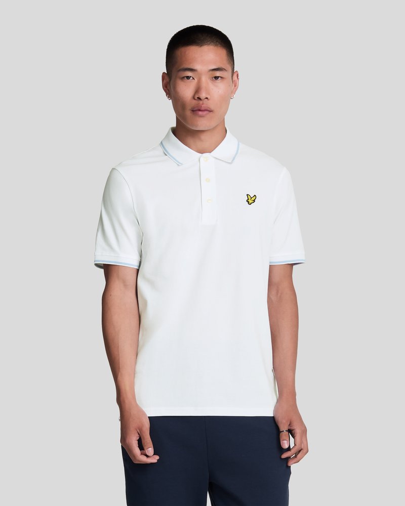 Hvit polo-skjorte med lyseblå detaljer på kragen og ermene; har et brodert gult fuglelogo på venstre bryst.