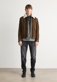 Chaqueta de ante marrón con un cuello de borrego blanco, cierre de cremallera, dos bolsillos frontales, camiseta gris, jeans oscuros y zapatos negros.