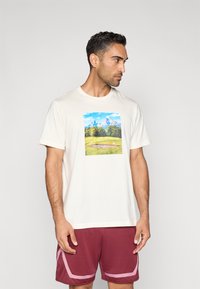 White cotton t-shirt featuring a vibrant landscape print of mountains and green fields. Paired with maroon athletic shorts with pink accents.