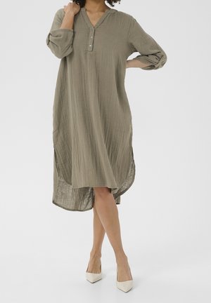 Donna che indossa un vestito ampio, verde oliva, a maniche lunghe abbottonato con maniche arrotolate e tacchi bianchi con punta appuntita, in piedi con le gambe incrociate.