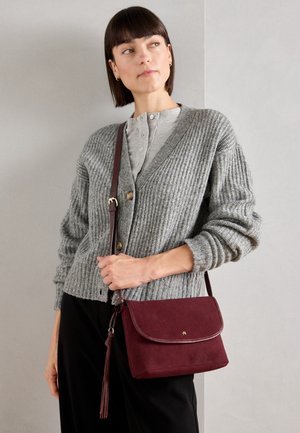 Kobieta o krótkich ciemnych włosach, ubrana w szary, zapinany na guziki kardigan i czarne spodnie, trzymająca bordową torebkę na ramię z frędzlami.