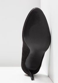 Schwarzer Wildleder-Highheel-Schuh, von der Sohle aus gezeigt, mit spitzem Zehenbereich und dünnem Absatz, der an einer weißen Ecke lehnt.