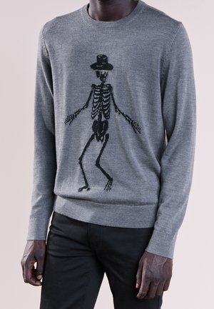 Mann trägt einen grauen Pullover mit einem schwarzen Skelett mit Zylinder auf der Vorderseite, kombiniert mit einer schwarzen Hose.
