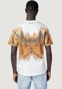 Man stands facing away wearing white T-shirt with orange and brown palm leaf print and light blue jeans against a plain background.