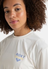 Camiseta deportiva blanca con un cuello redondo y mangas cortas. Presenta el logo "ROXY" en azul y amarillo claro en el pecho. Textura suave.