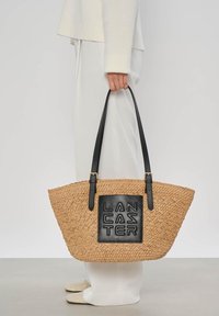 Personne en tenue blanche tenant un grand sac fourre-tout tissé beige avec des poignées noires et un patch carré noir portant l'inscription "LANCASTER".