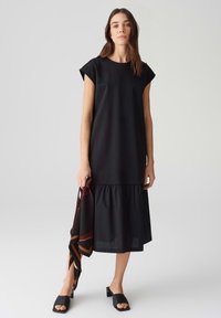Schwarzes Kleid mit kurzen Ärmeln und gestuftem Taillenbereich. Glatte Textur und knöchellang. Hält einen gemusterten Schal und trägt schwarze Rutschsandalen.