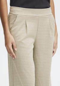 Pantalons à motifs beiges avec taille haute, comportant des plis, des poches latérales et un tissu texturé. Main positionnée sur la ceinture.