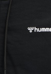Pantalon de sport noir avec une texture lisse, une fermeture éclair sur le côté et un logo "hummel" blanc. Le tissu présente un motif classique en maille.