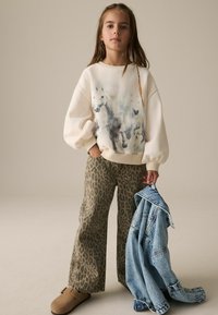 Kremna majica s grafiko konja, vzorčaste bež široke hlače in svetle jeans jakne, ki je draped over arm, v kombinaciji s rjavimi klompi.