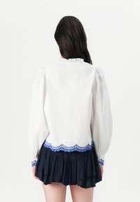 Blusa bianca con maniche lunghe a palloncino e colletto arricciato, con bordo ricamato a onde blu, abbinata a una gonna a strati blu scuro.