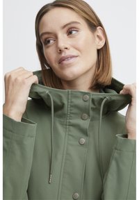 Veste imperméable verte avec un col haut, un cordon de serrage et des boutons pression. Présente une texture lisse et une coupe décontractée.