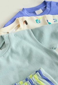 Tre børnetøjsbluser lagt oven på hinanden: en lyseblå bluse med lange ærmer, en cremefarvet bluse med farverige mønstre, og en blød lilla bluse. Alle har trykknapper.