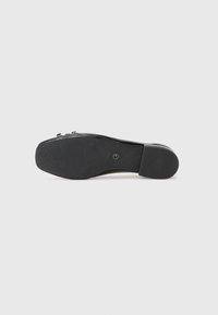 Semelle de chaussure noire avec une surface en caoutchouc texturée, présentant une légère courbure au niveau du talon et une petite empreinte de logo près du centre.