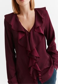 Blouse bordeaux avec un décolleté en V profond et des volants. Manches longues, tissu doux et accents de boutons en bas pour une coupe ajustée.