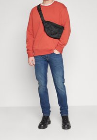 Bolso cruzado negro de nailon con múltiples bolsillos, llevado sobre un sudadera de color óxido, combinado con unos jeans de mezclilla azules y botas negras.