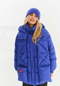 Modrá prošívaná bunda s vysokým límcem, velkými kapsami a diagonálním prošitím. Doprovázena odpovídající fialovou pletenou čepicí.