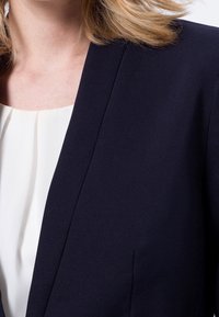Tmavě modrý blejzr s přizpůsobeným střihem, který má klopový límec a hladkou texturu. Pod ním se nachází bílá plisovaná blůza.