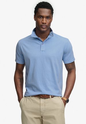 Mann steht mit den Händen in den Taschen, trägt ein hellblaues Poloshirt, beige Hose, einen braunen geflochtenen Gürtel und eine Armbanduhr.