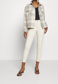 Žena v béžovém kostkovaném saku přes černý top, v světle krémových úzkých kalhotách a černých sandálech na podpatku s pásky, stojící na bílém pozadí.