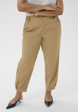 Persoon draagt beige taps toelopende broek met elastische zoom, wit topje, zwarte puntige slingback-hakken en gouden ring en armband.