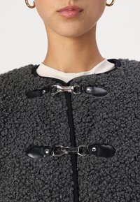 Manteau gris texturé en matériau flou et bouclé. Présente des accents en cuir noir et une quincaillerie en métal argenté pour la fermeture. Encolure ronde.