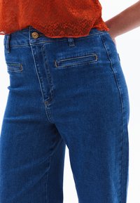 Modré džíny z denimu s klasickým střihem, předními kapsami, jedním knoflíkovým zapínáním a kontrastními švy.