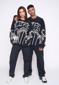 Pulovere negre tricotate cu desene abstracte albe, asortate cu pantaloni cargo negri și adidași. Textură netedă, croială lejeră și detalii suprapuse.