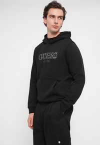 Mężczyzna w czarnej bluzie Guess z kieszenią 'kangur' i czarnych spodniach cargo, stojący na tle białym, patrząc na bok.