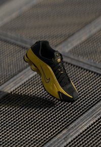 Μαύρο και κίτρινο αθλητικό παπούτσι Nike για τρέξιμο με σχέδιο κορδονιών, τοποθετημένο σε μεταλλική σχάρα με διαγώνιες σκιές.