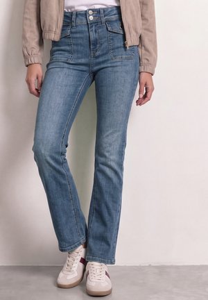Femme portant un jean bleu taille haute à jambes droites, des baskets blanches avec des bandes bordeaux, une veste beige et un haut blanc sur un fond uni.