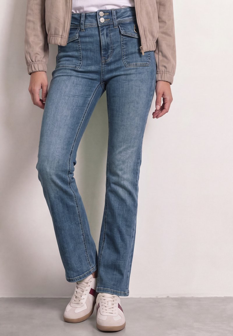 Femme portant un jean bleu taille haute à jambes droites, des baskets blanches avec des bandes bordeaux, une veste beige et un haut blanc sur un fond uni.