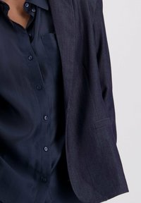 Mörk marinblå kavaj med skarpa slag, mjuk textur och skräddarsydd passform, bärs över en matchande skjorta med knappar och bröstficka.