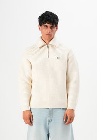 Young man wearing cream knit half-zip sweater and light blue jeans, standing against a plain white background.