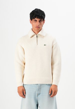 Young man wearing cream knit half-zip sweater and light blue jeans, standing against a plain white background.