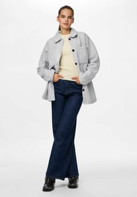 Veste en mélange de laine grise avec un col structuré, des poches avant et des boutons noirs. Associée à un jean large bleu foncé et des chaussures noires.